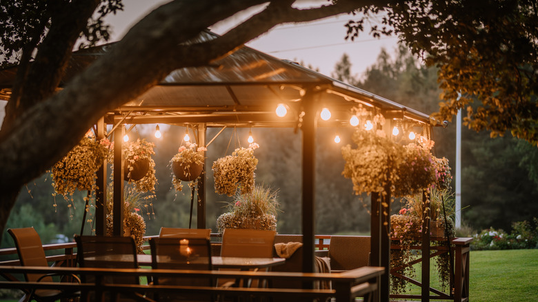 A gazebo outside with hanging plants, dining seating, and strung lights at dusk