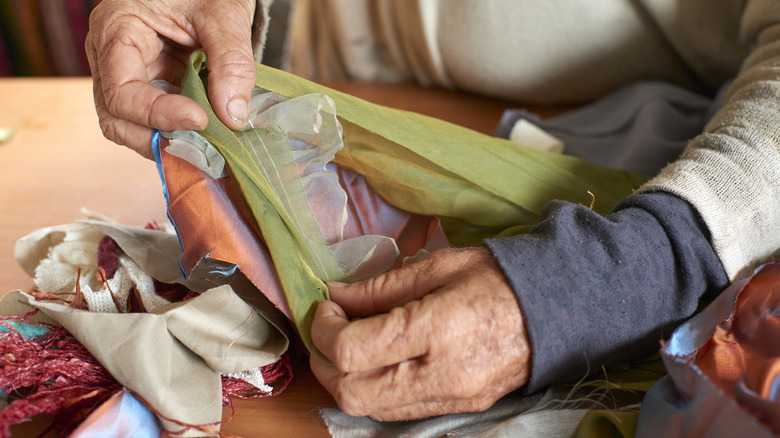 A pair of mature light-skinned hands sort through strips of colorful fabric on a table top.