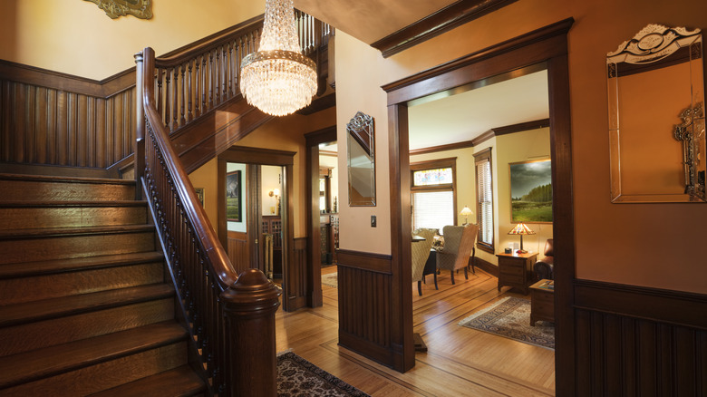 Foyer of a Victorian home with staircase, crystal chandelier, and a view into a living room.
