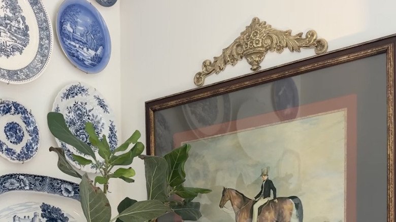 A brass wall hanging mounted above a framed painting with plates on the wall and a houseplant in front.