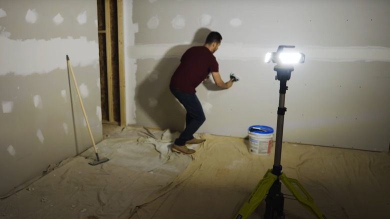 Man mudding drywall in a dark room with light from a Ryobi tripod light