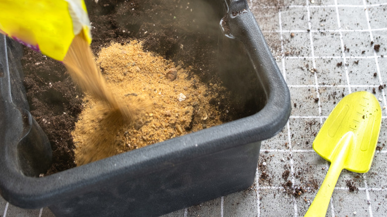 A person pours horticultural sand on top of some potting mix in a rectangular black plastic planter with a yellow garden trowel nearby.