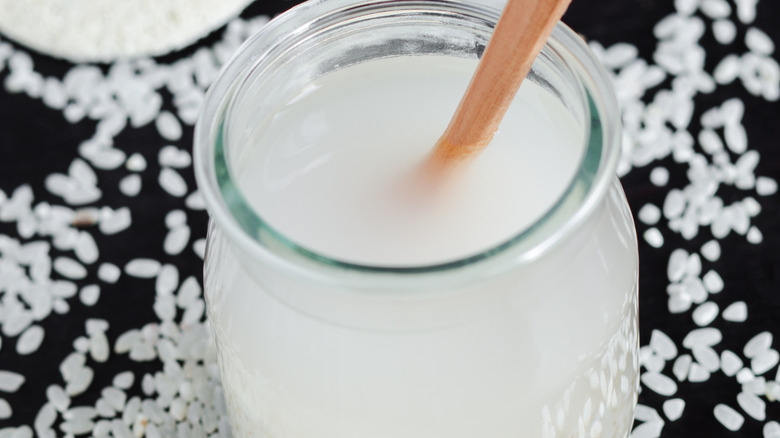 glass jar full of rice water with a wood spoon in it on a dark surface covered in dry rice