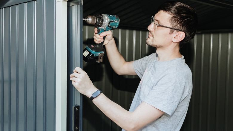 Man assembling a gray metal storage shed with a drill.