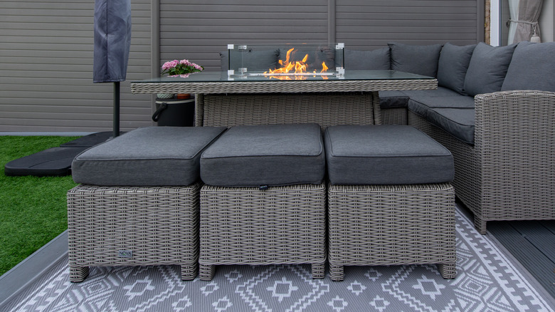 A patio with dark grey rattan furniture and pillows with a table fire and a patterned rug.