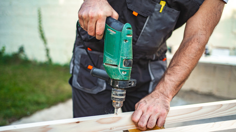 A male carpenter using electric drill on a wooden plank outdoors.