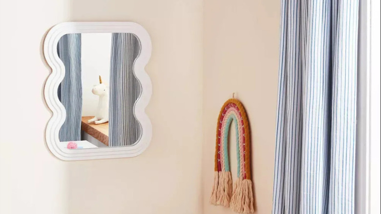 Target kid's mirror with a white wavy frame on a beige wall with a piece of rainbow rope decor and blue striped curtains