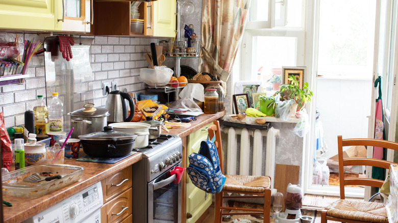 A cluttered kitchen with pans, jars, and dishes on counter and stove top.