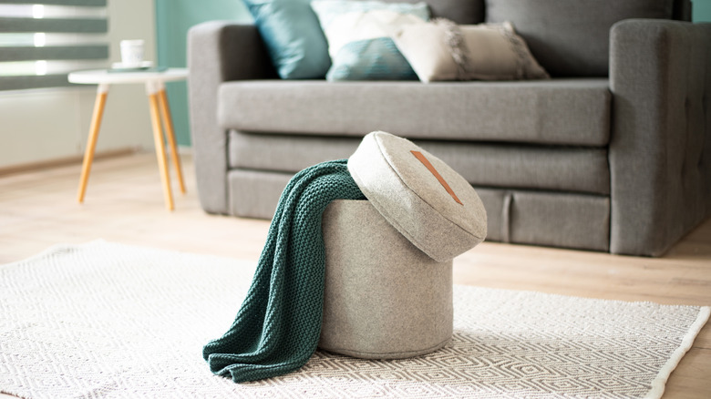 Felt storage ottoman with green knitted throw on geometric rug in a bright Scandinavian minimalist living room, featuring soft gray sofa, decorative pillows and natural light atmosphere