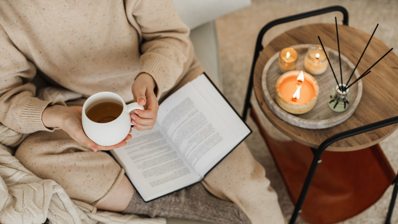 cozy fall set up with candles on side table while woman drinks tea and reads
