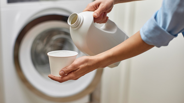 A woman's hands are showing pour bleach in front of a washing machine