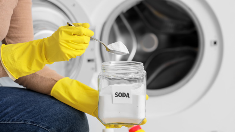 A woman with yellow gloves is spooning washing soda in front of a washing machine