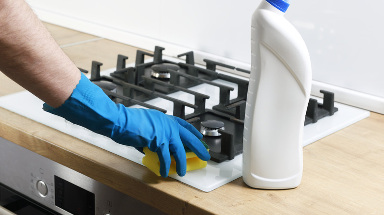 A gloved hand cleaning a stove top with bleach and a sponge.