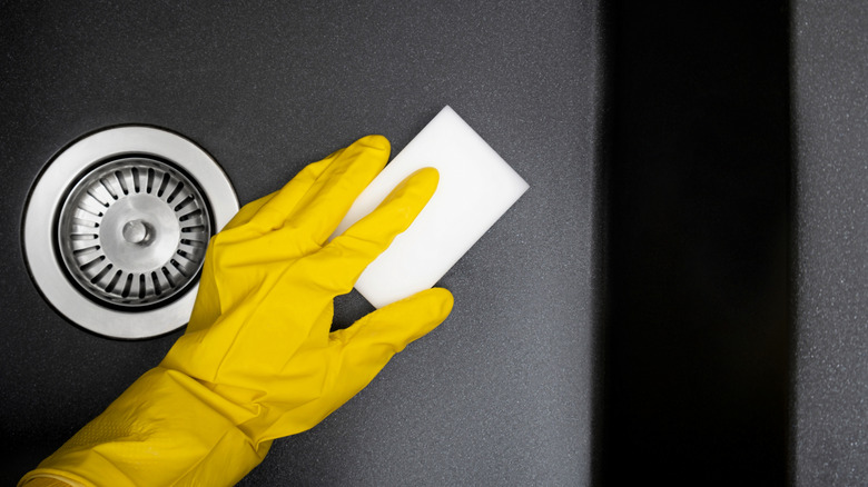A gloved hand using a melamine foam sponge to clean the bottom of a kitchen sink.