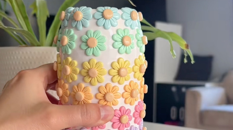 A hand holding a vase covered in clay flowers in a variety of spring colors with a potted plant in the background