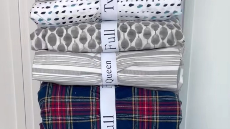 Patterned bedsheets in whites, grays, stripes, and red and blue plaid are bundled with white elastic bands that state the sizes of the sheets.
