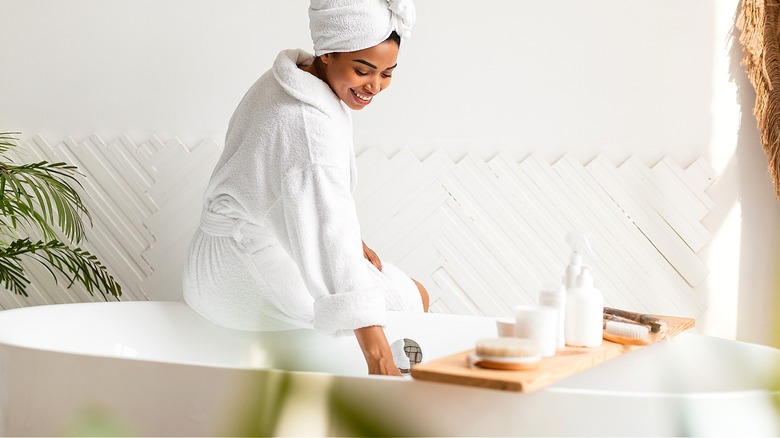 A person smiles as they sit on the edge of a tub and prepare a bath