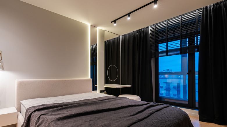 Dim bedroom with track lighting, partially made bed, and large windows with black out curtains.