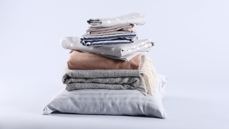 A neatly stacked pile of folded bedding — including pillows, blankets, and patterned sheets in soft gray, white, and beige tones — is set against a plain light background.
