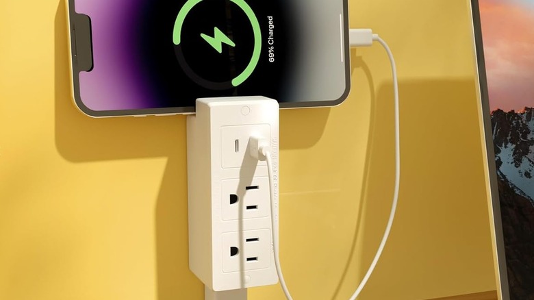 A white electrical outlet extender behind a table holding and charging a smart phone
