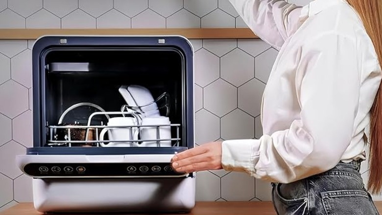 Woman holding door of a small white dishwasher on a countertop