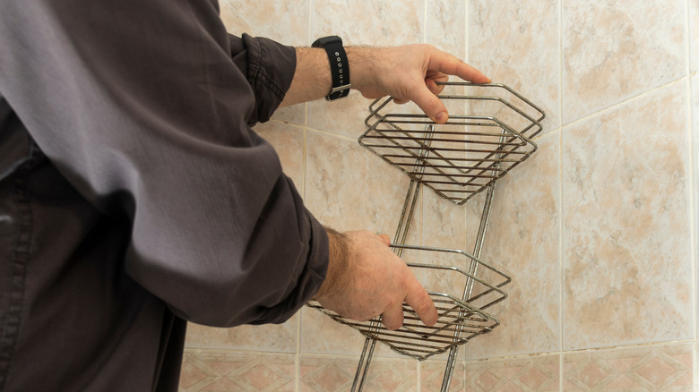 A person removing an old, rusted shower caddy from the bathroom wall
