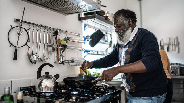 A smiling senior Black man cooks in a wok over a gas stove.