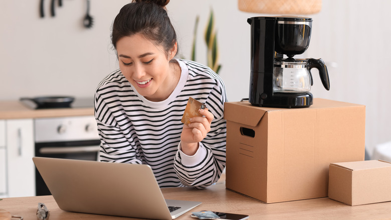 A smiling person using the laptop for online shopping along with a credit card. On the bench is also a coffee maker on a cardboard box