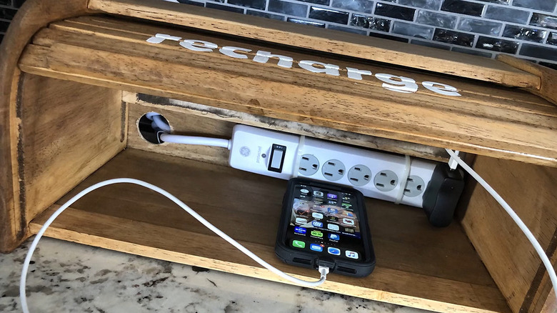 Wooden bread bin with white lettering "recharge" and a surge protector and phone plugged in