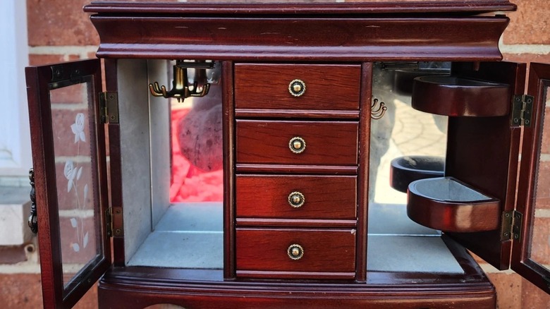 Large vintage wooden jewelry box shaped like an armoire with opened glass doors in front of a brick wall