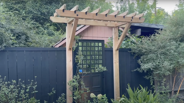 A wooden arbor frames a door and mirror on a backyard garden fence.