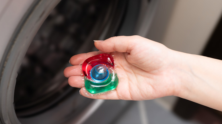 A hand holding a detergent pod in front of an open washing machine