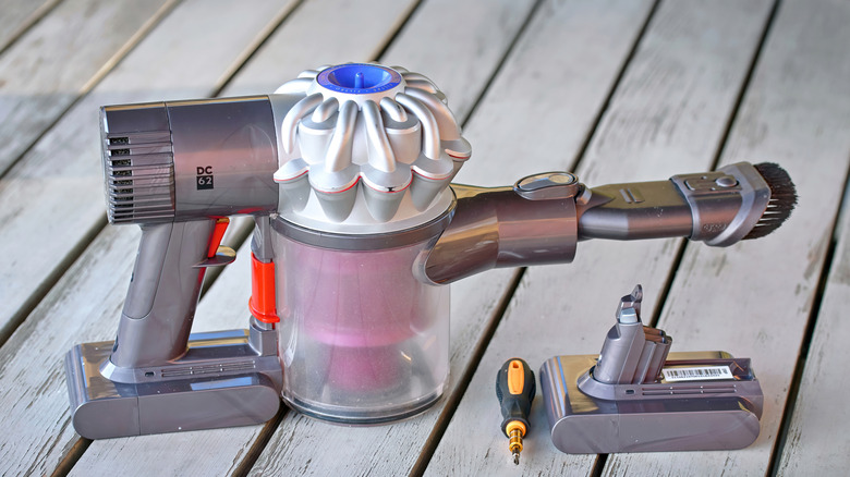 A handheld Dyson vacuum, screwdriver, and replacement battery sit on a wood deck