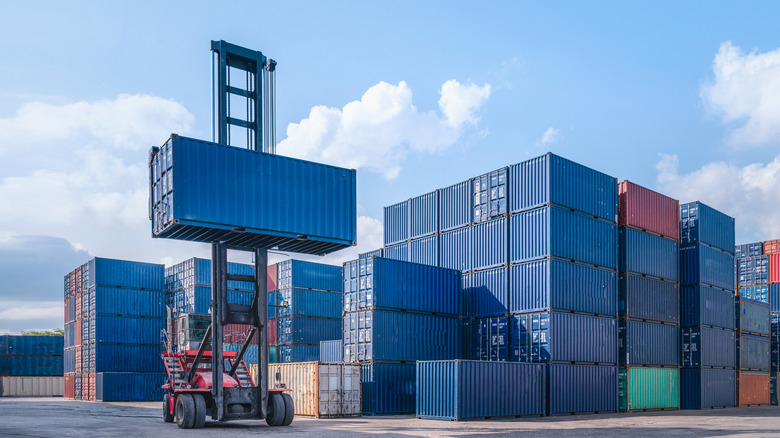 Blue shipping container being lifted by machine next to a stack of other containers in shipyard