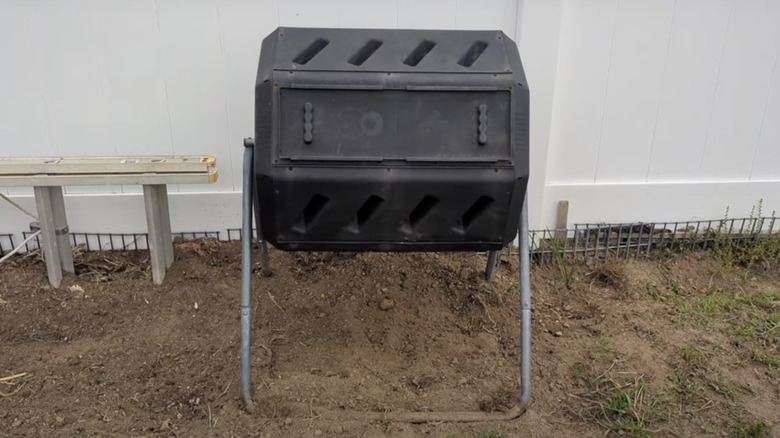 A black plastic tumbling composter with two compartments on a metal stand in a backyard.