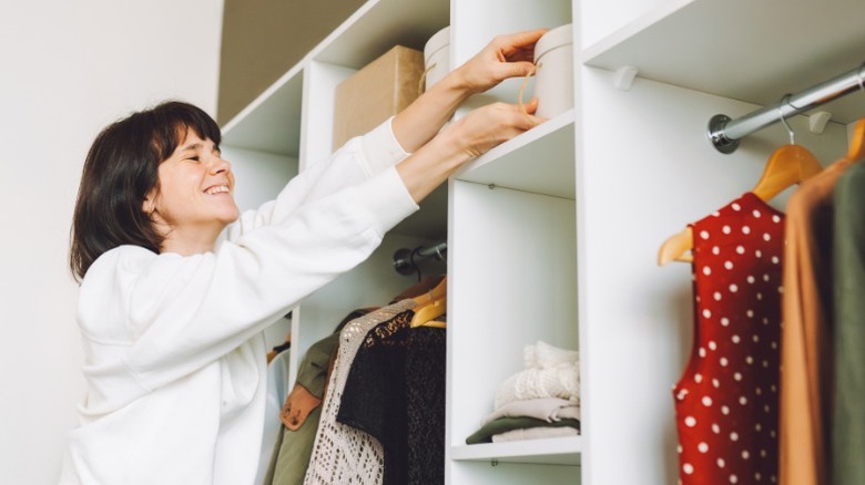 smiling woman reaching for container on bedroom closet shelf