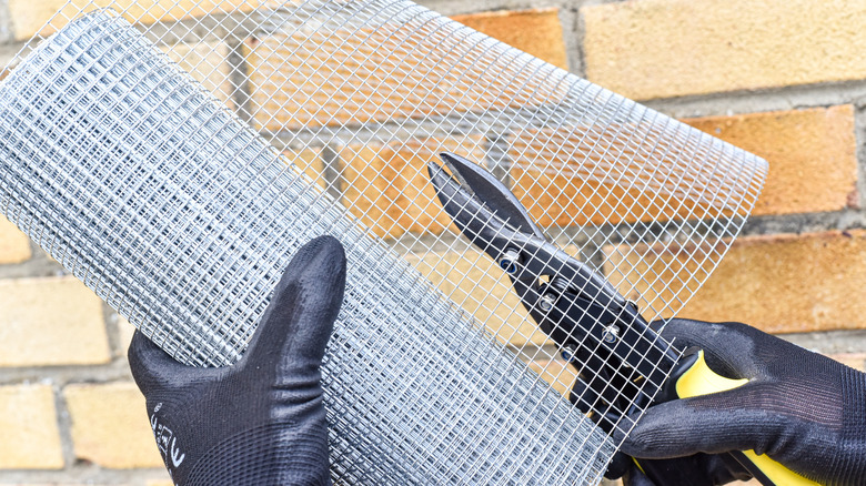 A pair of gloved hands cuts a roll of square wire mesh using wire cutters.