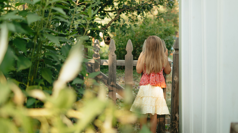 A young girl in a red checkered top and yellow skirt stands at a wooden fence in a lush garden, looking away.