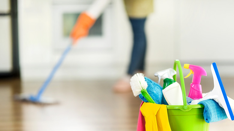 A bucket with spray bottles and other cleaning supplies is in the foreground as a person cleans a wood floor in a blurred background