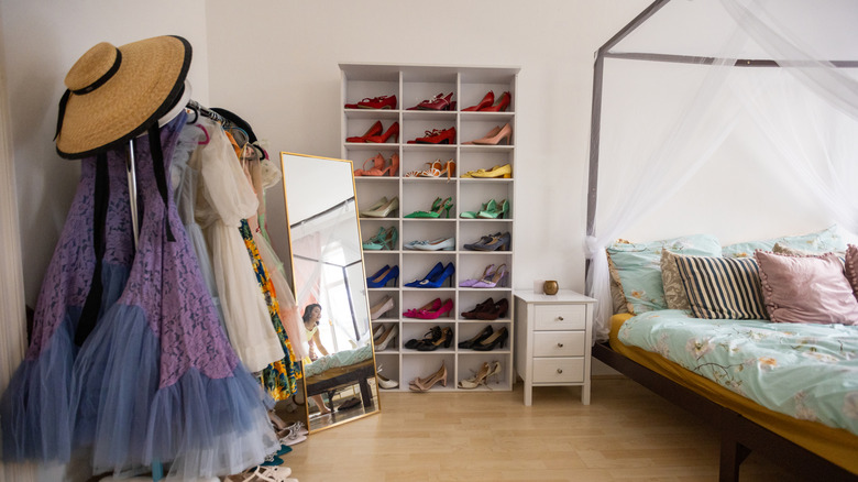 A collection of colorful shoes in an organizer near a bed, nightstand, mirror, and clothing rack