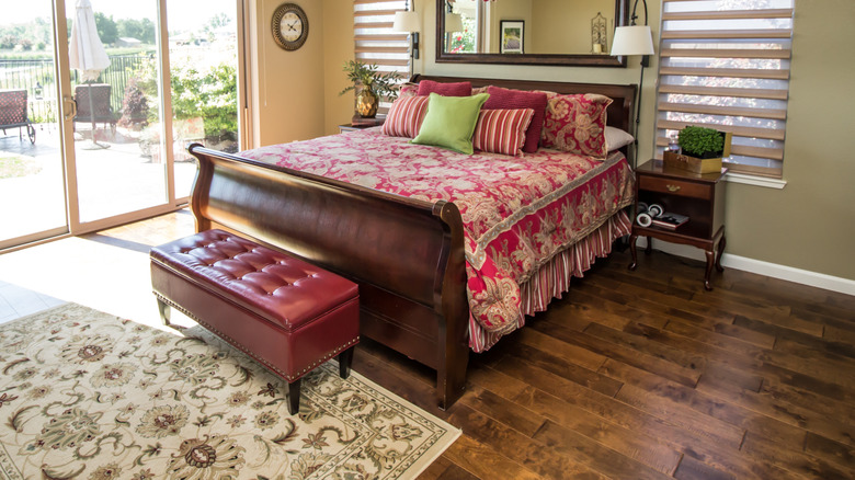 A sunny bedroom featuring a bed with patterned covers and pillows, a ceiling fan with light, and hardwood flooring with a carpet.