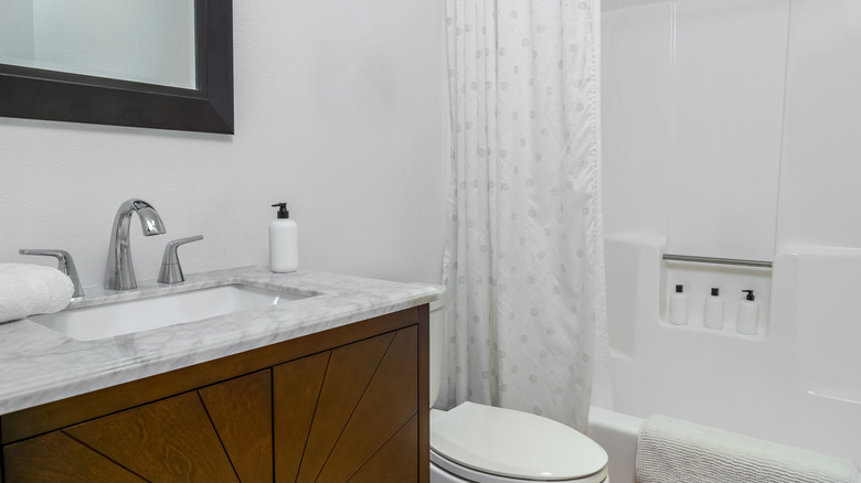 A modern bathroom with a white marble and wood vanity and white toilet with a tub in the background