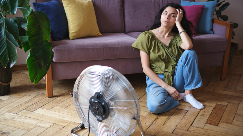 Tired exhausted young woman sitting on floor in front of electric fan, cooling down refreshing during heatwave.