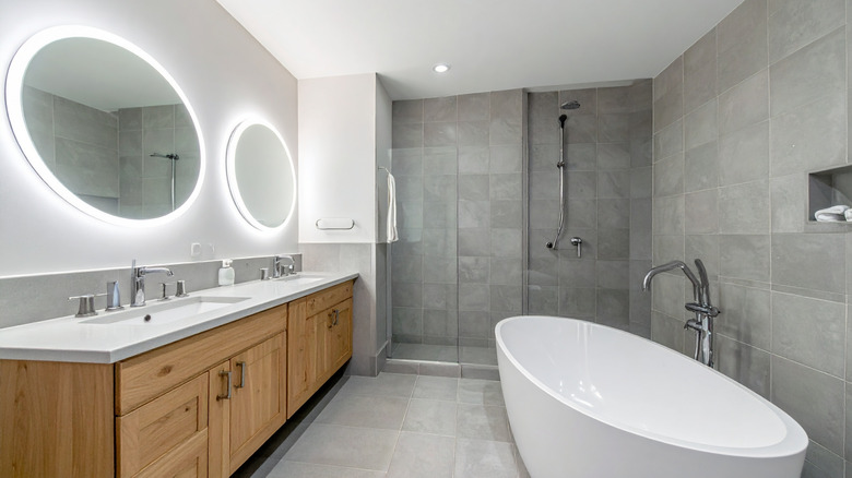 A contemporary bathroom features gray tile walls, a freestanding bathtub, double vanity with illuminated mirrors, and a glassenclosed shower.