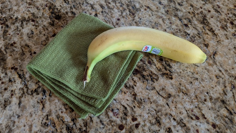 A banana in its peel and a green microfiber cloth lie on a granite countertop.