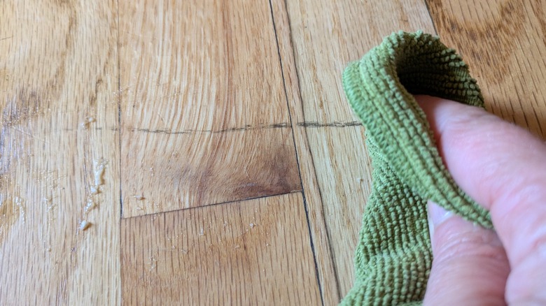 A light skinned hand rubs a scuffed wood floor with a cloth.