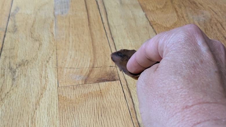 A light-skinned hand rubs a piece of brown banana peel on a floor scuff.