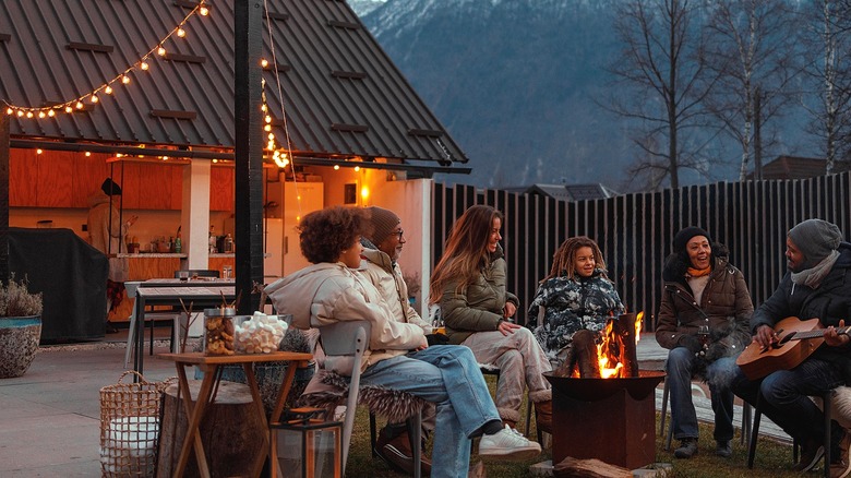 People wearing cold-weather clothing sit around a fire with a string of lights seen in the background near a kitchen