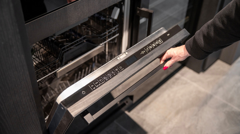 Woman in dark clothing opens the empty dishwasher door with a manicured hand featuring red nail polish