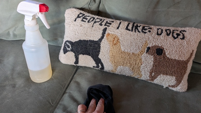 A spray bottle of DIY cleaner sits on a couch, and a hand with a rag rubs the upholstery.